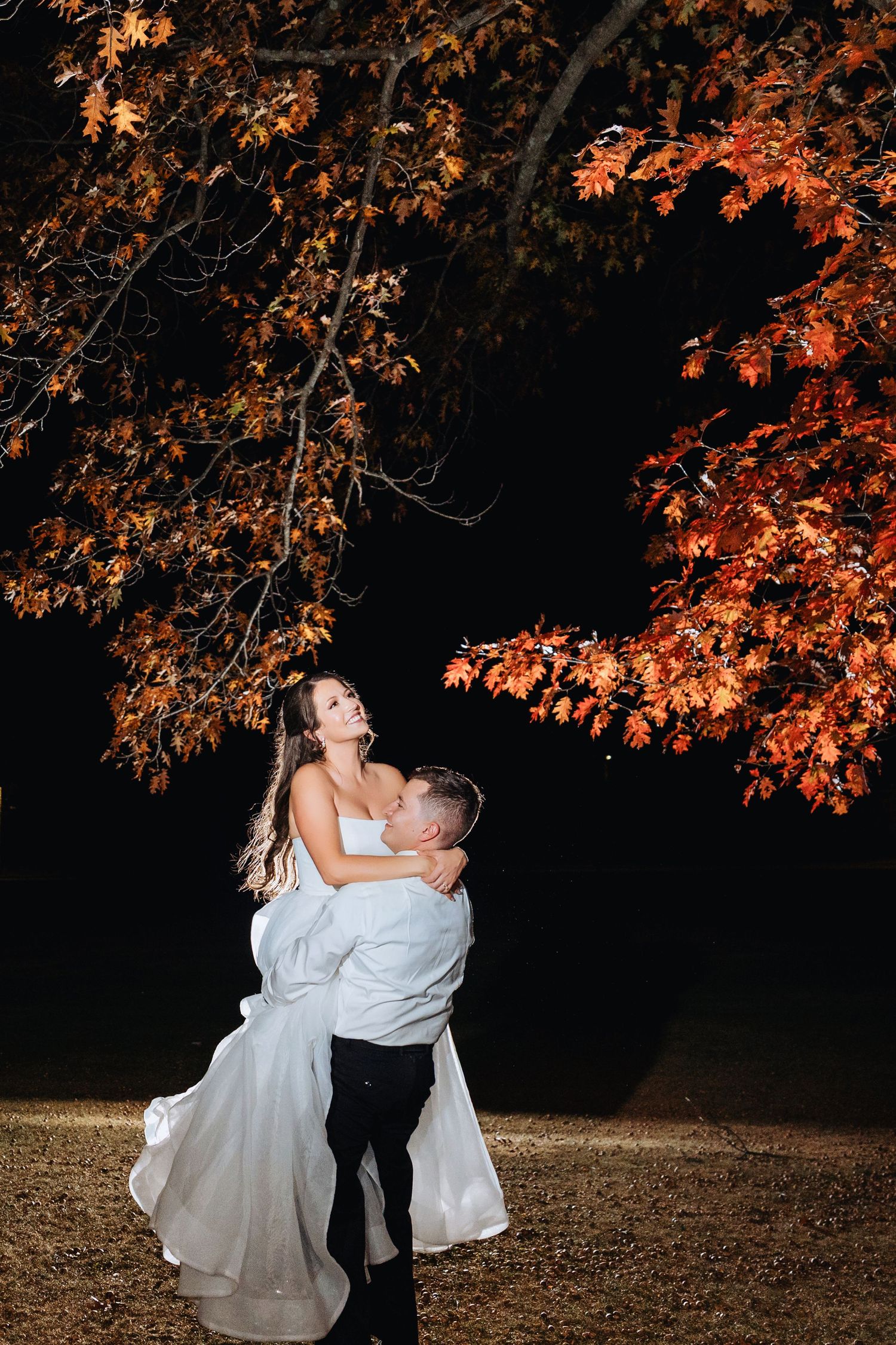 Romantic couple embraces under vibrant autumn maple trees with orange leaves against dark evening sky.