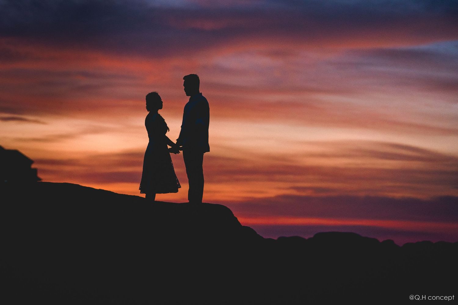 Two silhouetted figures stand together against a dramatic orange and purple sunset sky.