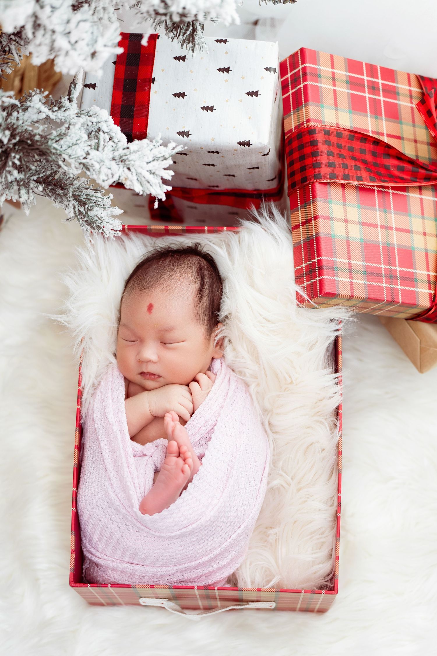 A sleeping newborn nestled in a white blanket inside a red plaid Christmas gift box.
