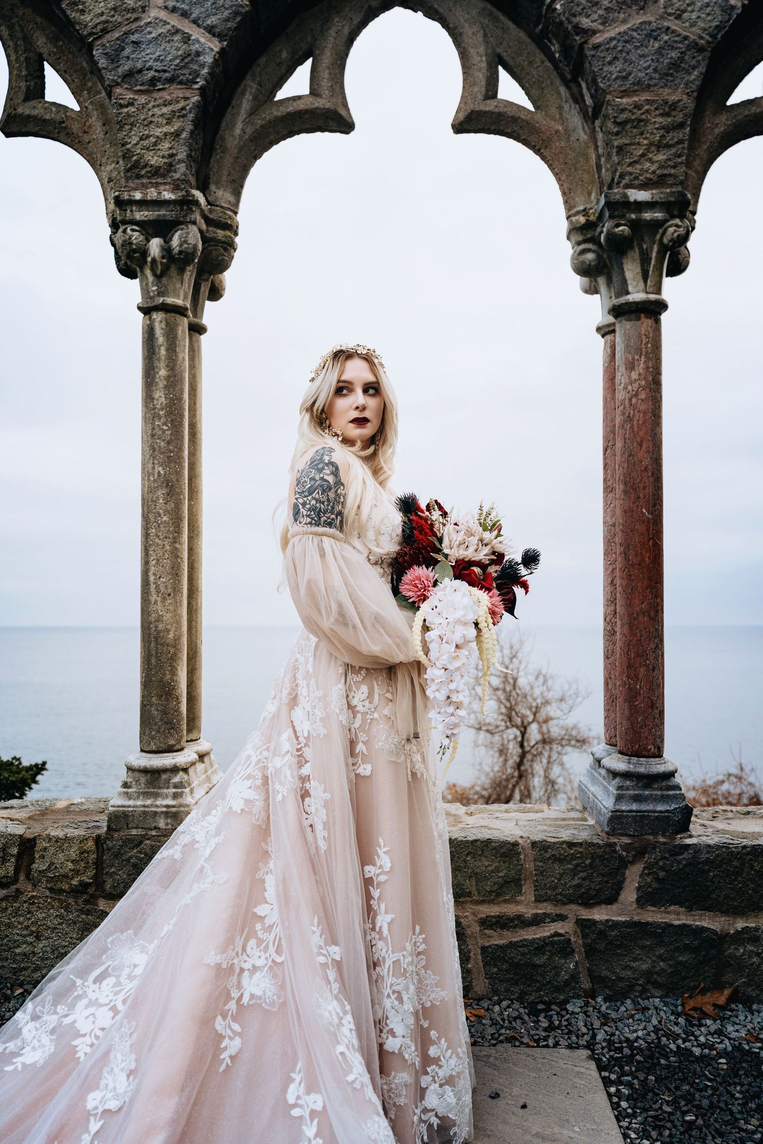 A person in a flowing blush pink lace wedding gown poses in an ornate stone archway against a cloudy sky.