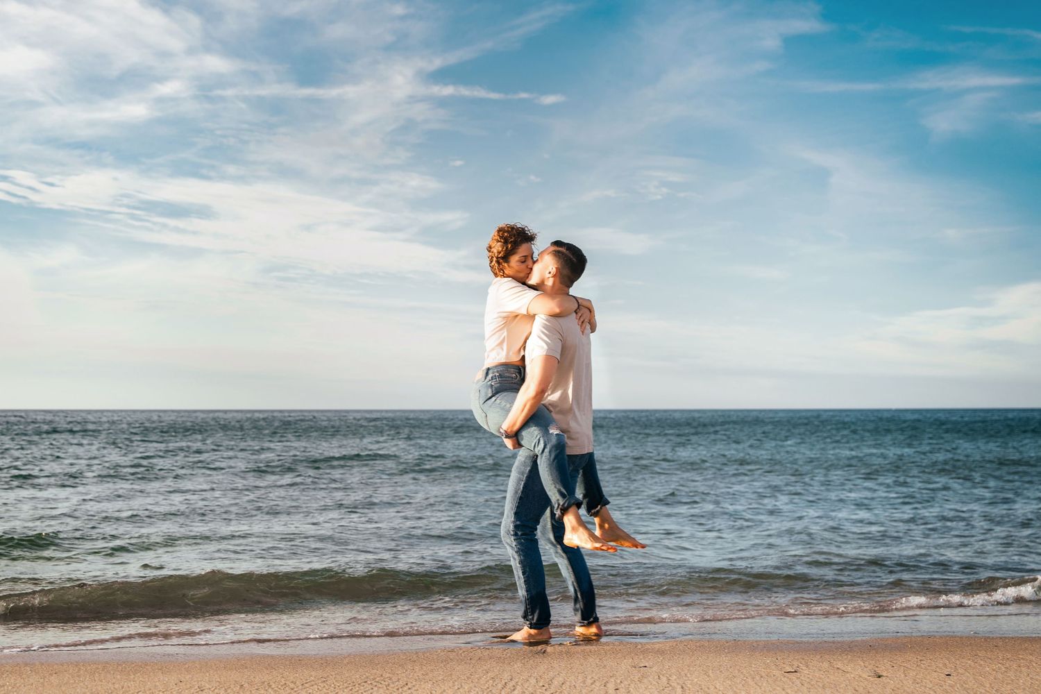 Romantic couple embracing and kissing on a sunny beach with blue ocean waves and cloudy sky in the background.