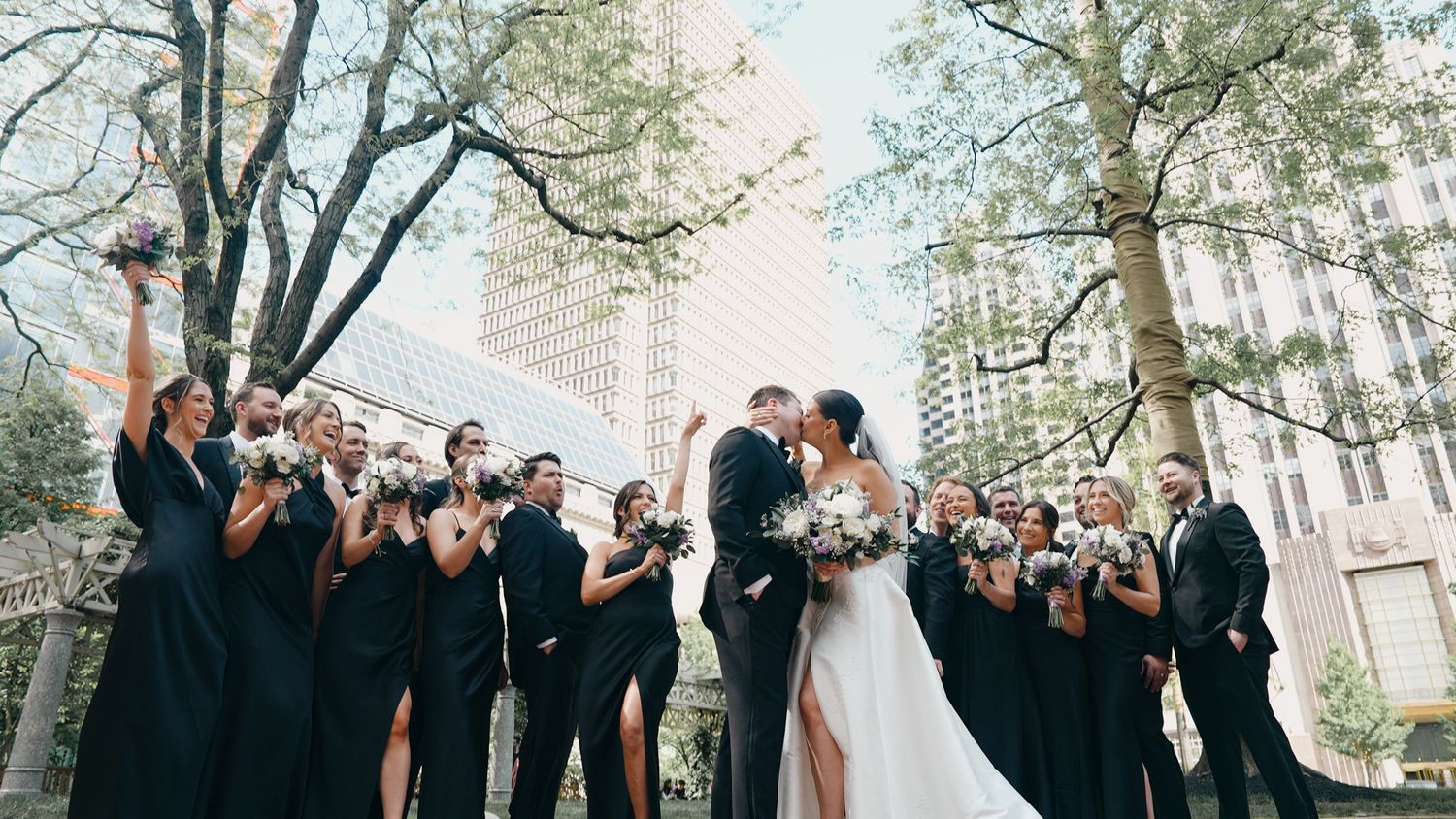 Wedding party in black attire celebrates with couple kissing under trees in an urban setting.