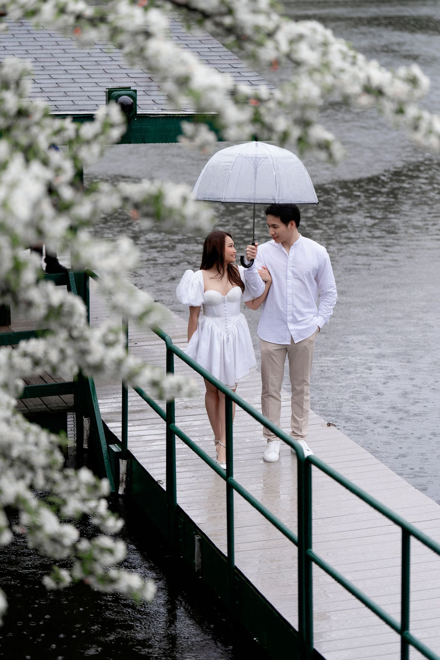 A couple in white outfits poses romantically on a dock near blooming cherry blossoms on a rainy spring day.