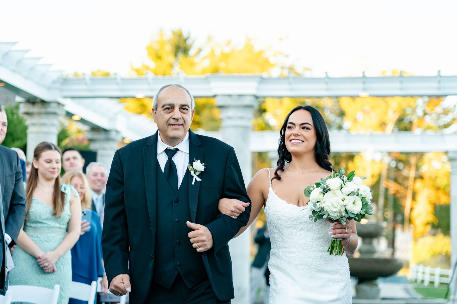 Wedding ceremony processional taking place outdoors during golden hour with autumn foliage.