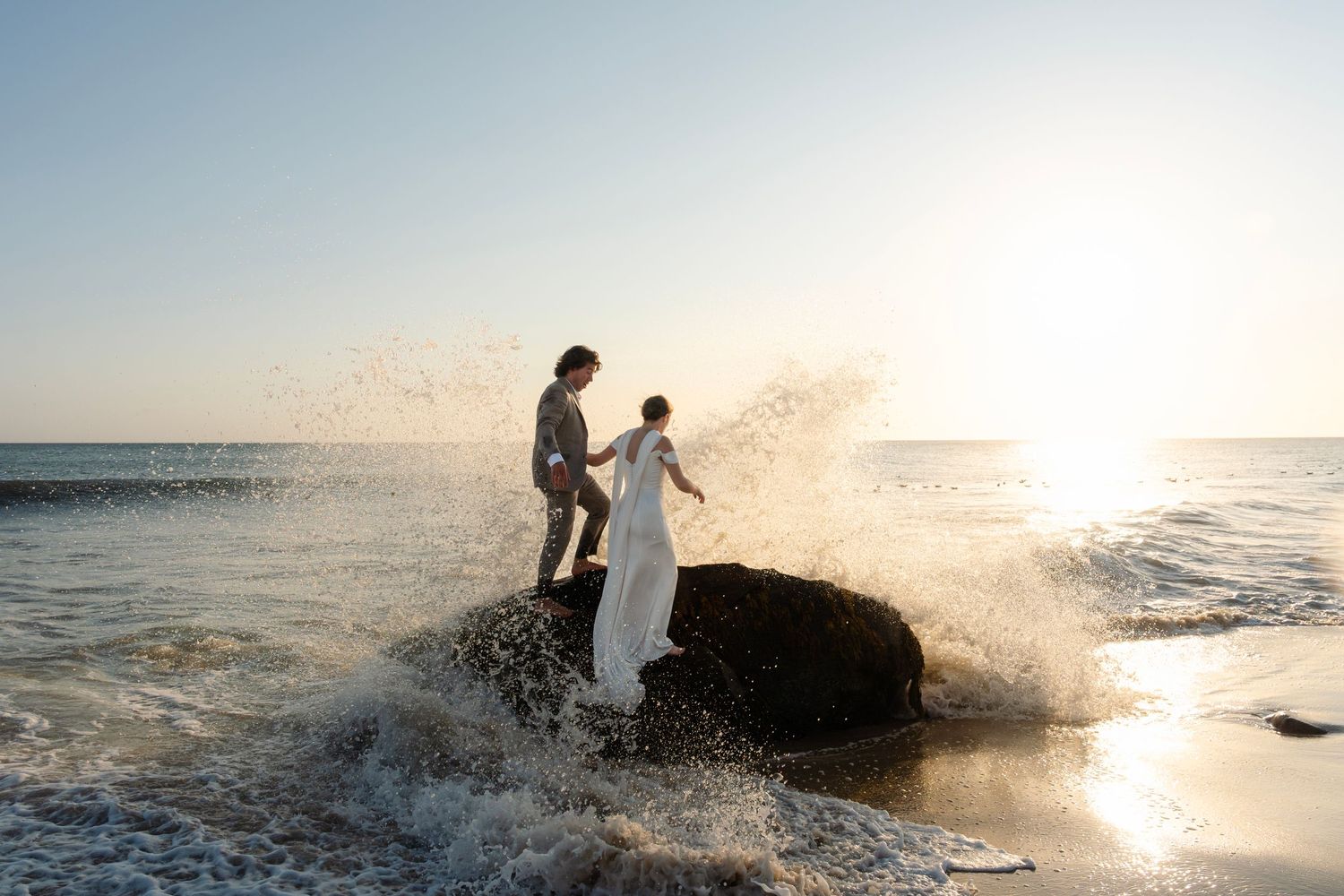 Couple sitting on rocks by ocean with waves splashing during golden hour.