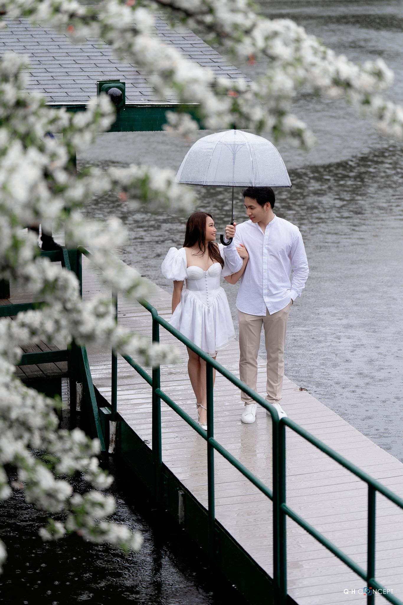 A couple in white outfits shares an umbrella while standing on a dock, with cherry blossoms framing the romantic scene.