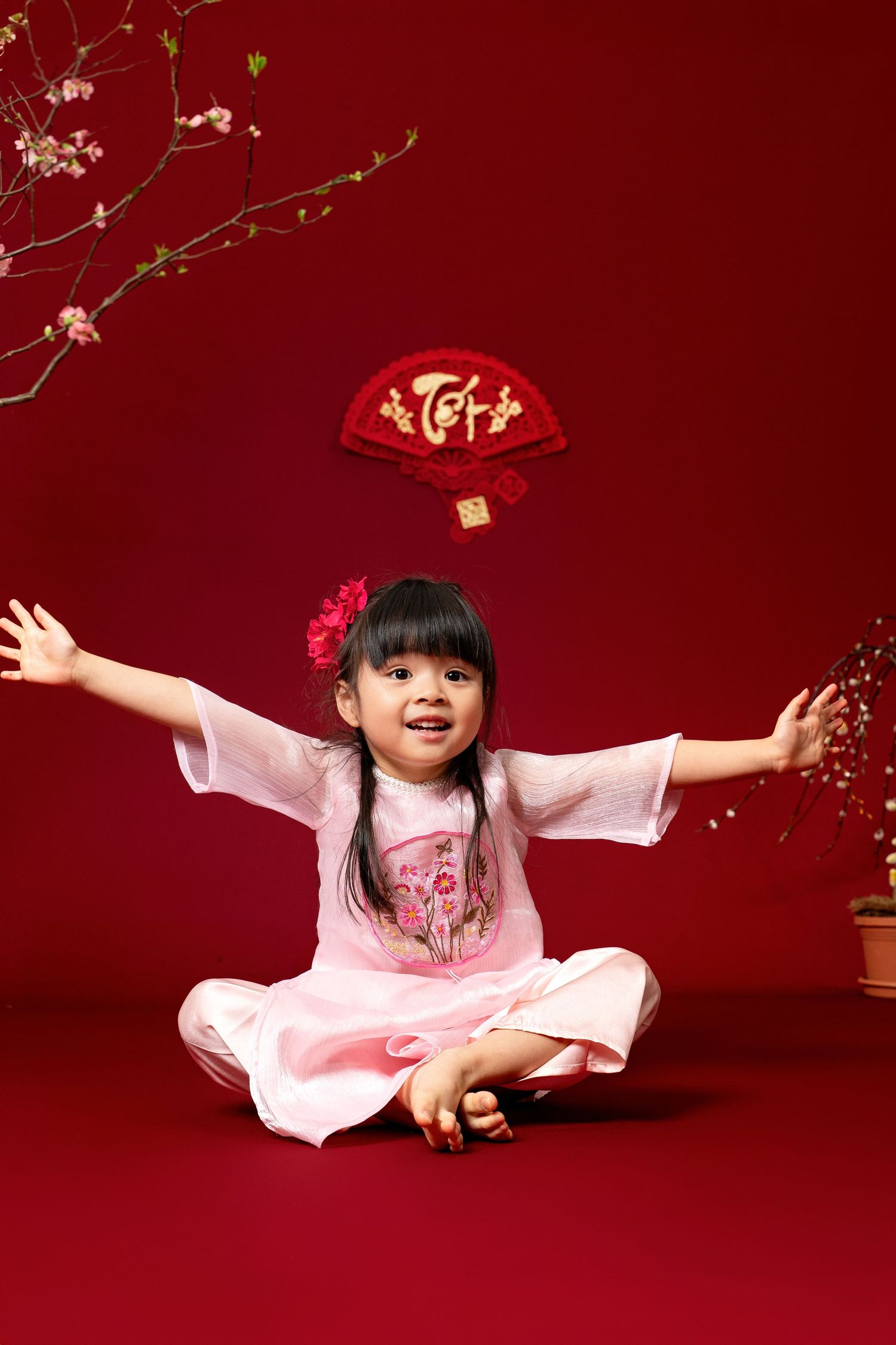 Child in pink dress plays with red lanterns and decorative elements against deep red backdrop during Chinese New Year celebration.