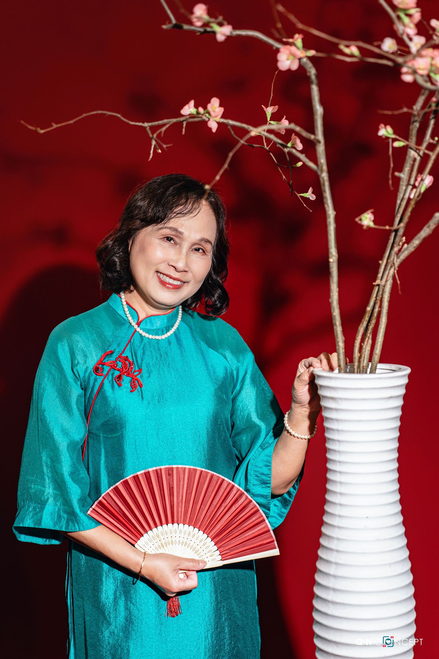 Traditional Asian-inspired fashion with turquoise qipao dress and red paper fan against deep red backdrop.