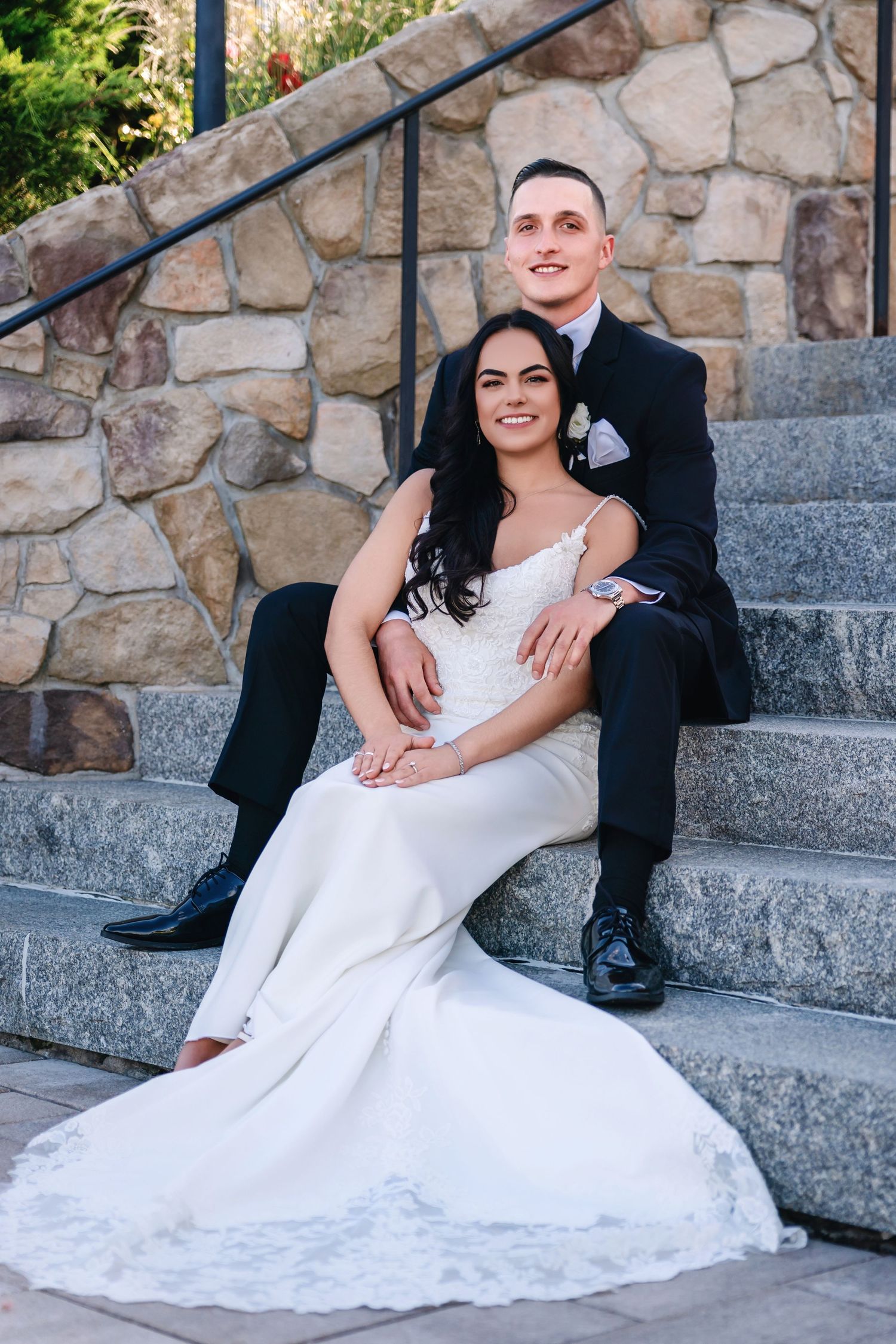 Wedding couple poses on stone steps, bride in white lace gown and groom in dark suit.