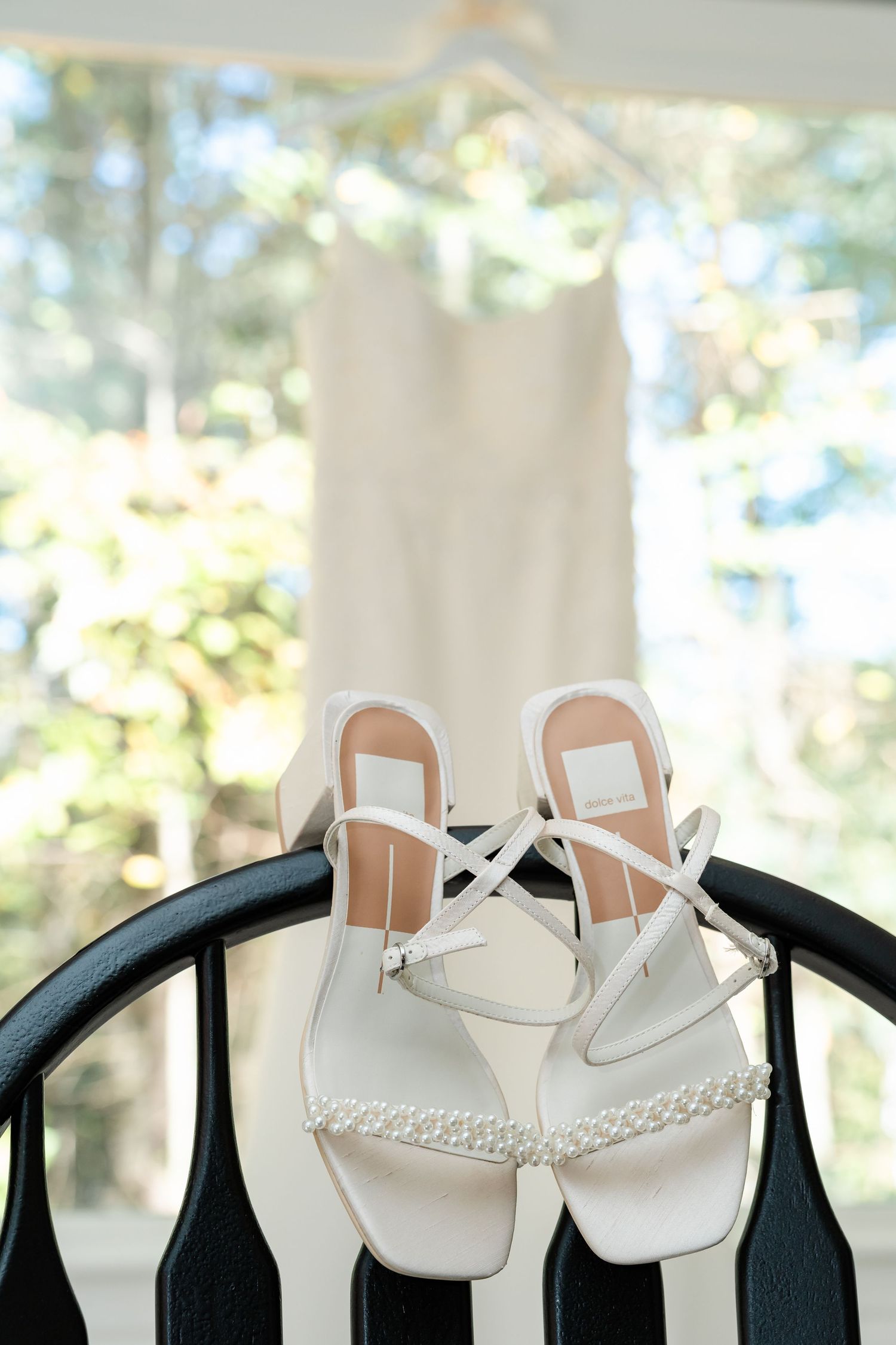 Row of white bridal sandals hanging on hangers against a bright window background.