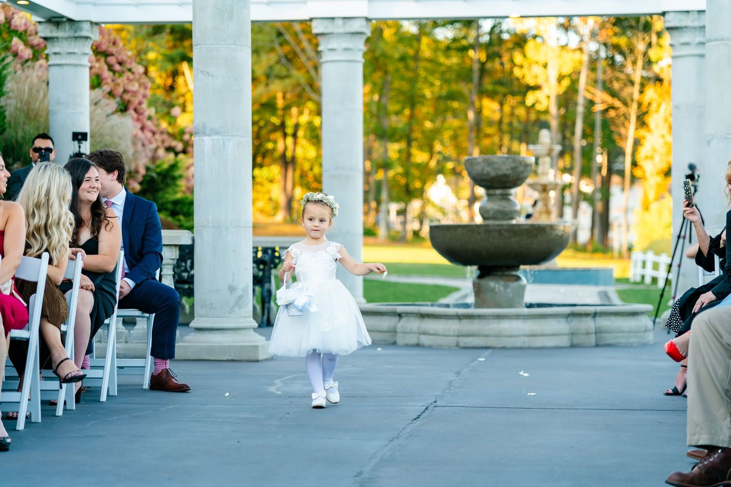Wedding ceremony guests gather at an elegant outdoor pavilion with white columns during golden hour.