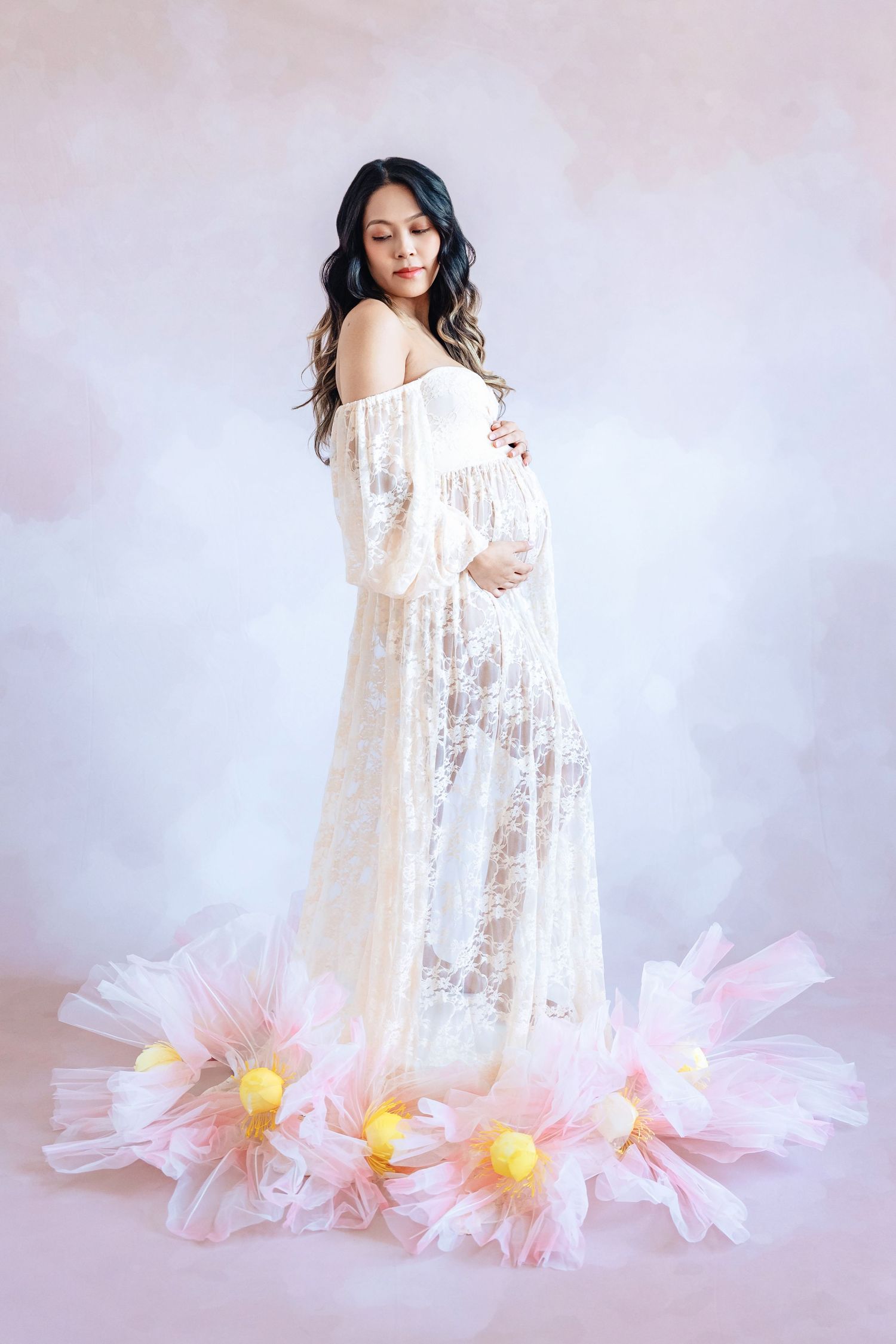 Ethereal maternity photos with flowing white lace dress surrounded by pink paper flowers on light background.