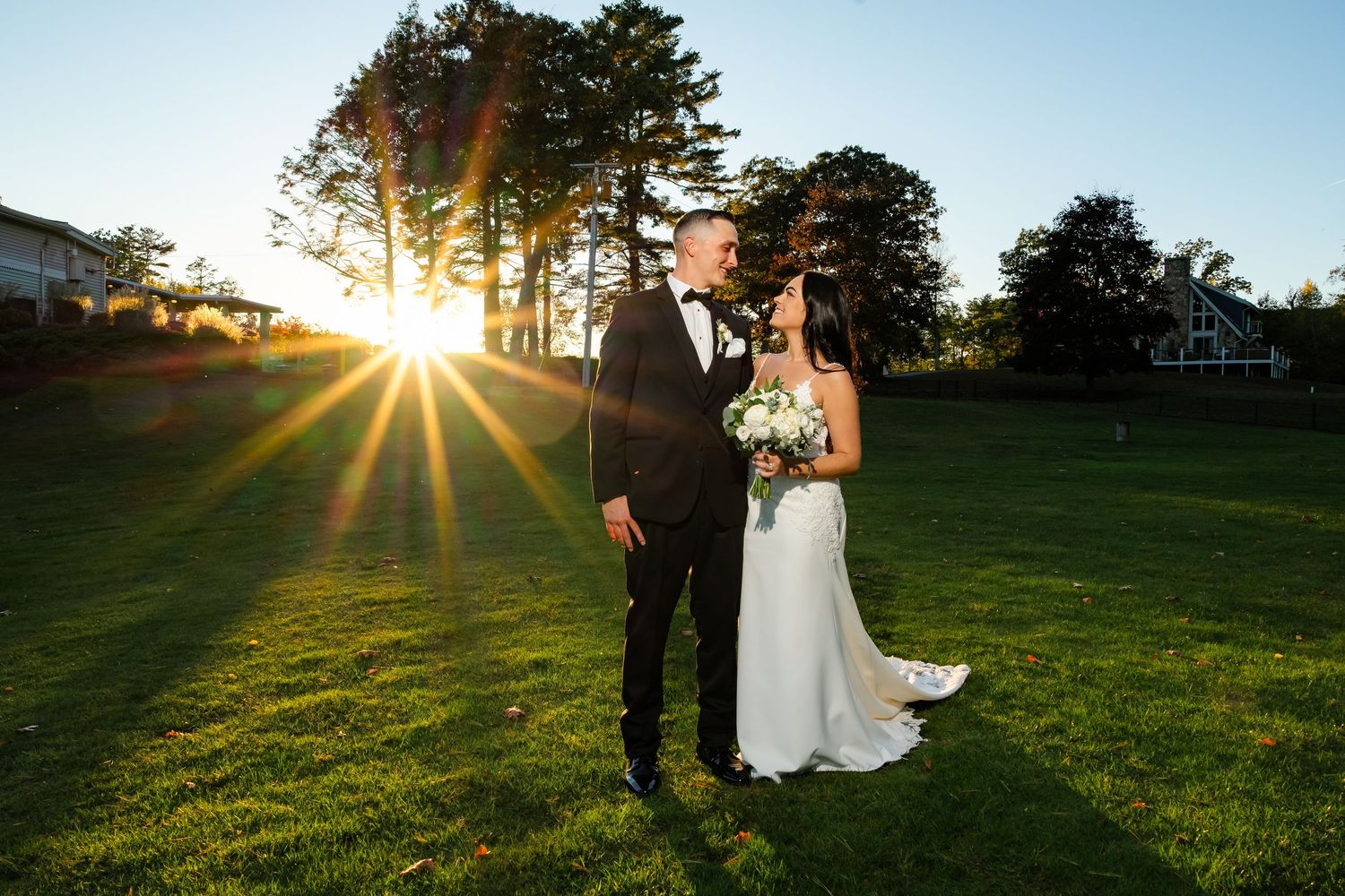 Golden sunlight streams through trees as a couple takes romantic sunset wedding photos in a grassy field.