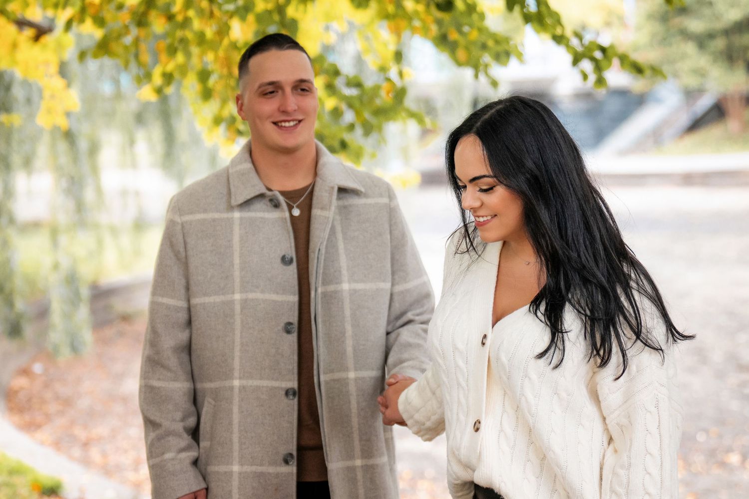 A couple in autumn attire shares joyful moments together in a park with golden fall foliage in the background.
