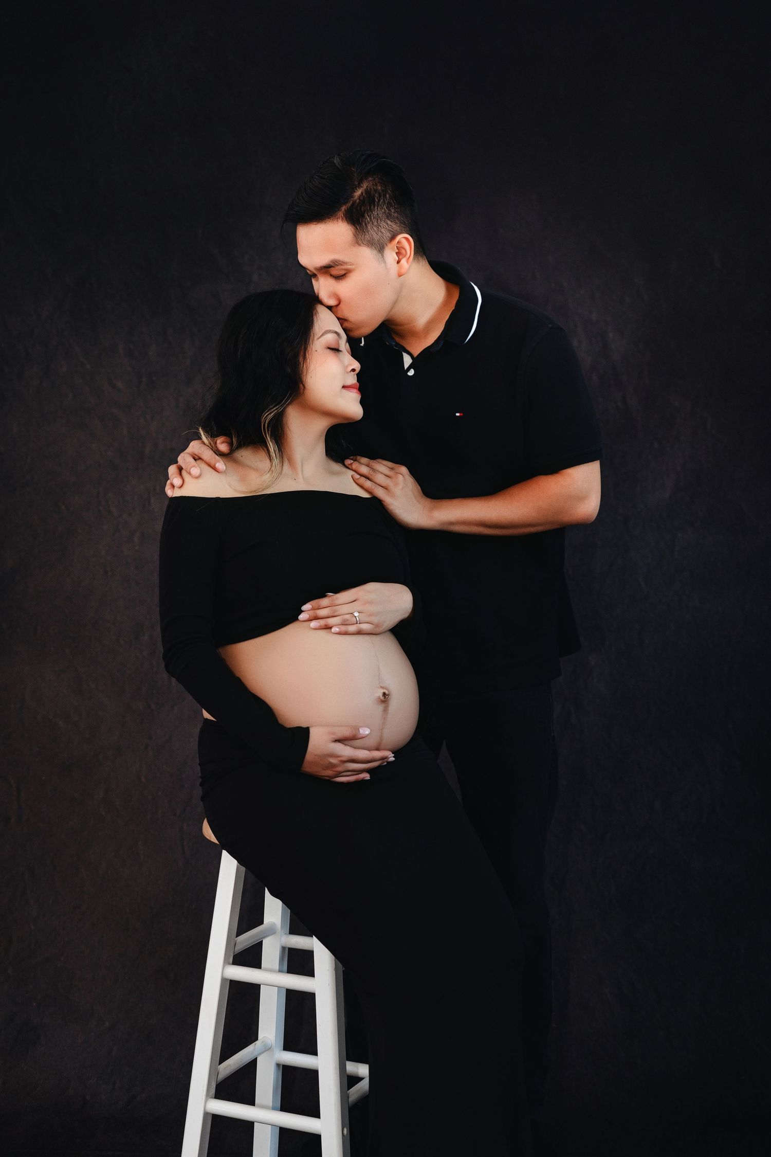 Intimate maternity portraits on white stool against dark background showing couple's connection.