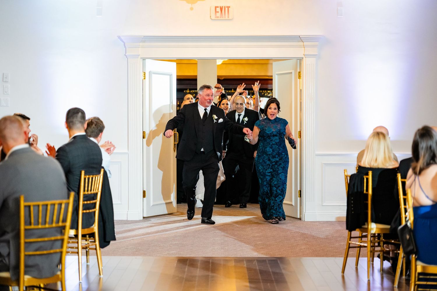 Wedding reception guests dancing in an elegant white ballroom with gold chiavari chairs along the perimeter.