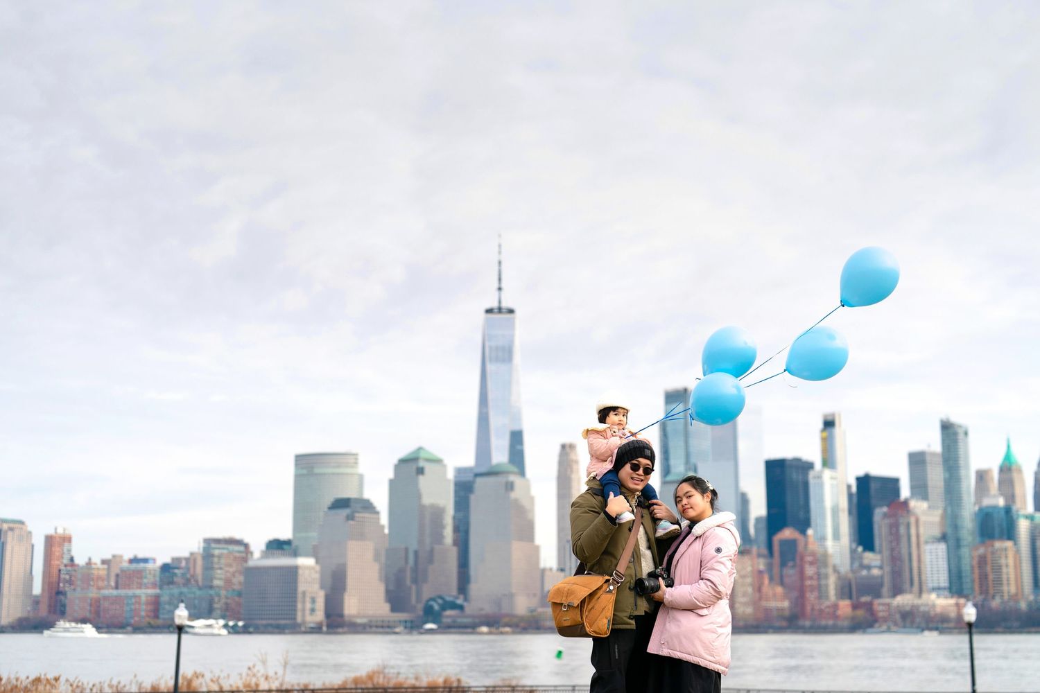 Family with blue balloons posing by the Hudson River with the New York City skyline and One World Trade Center in the background.