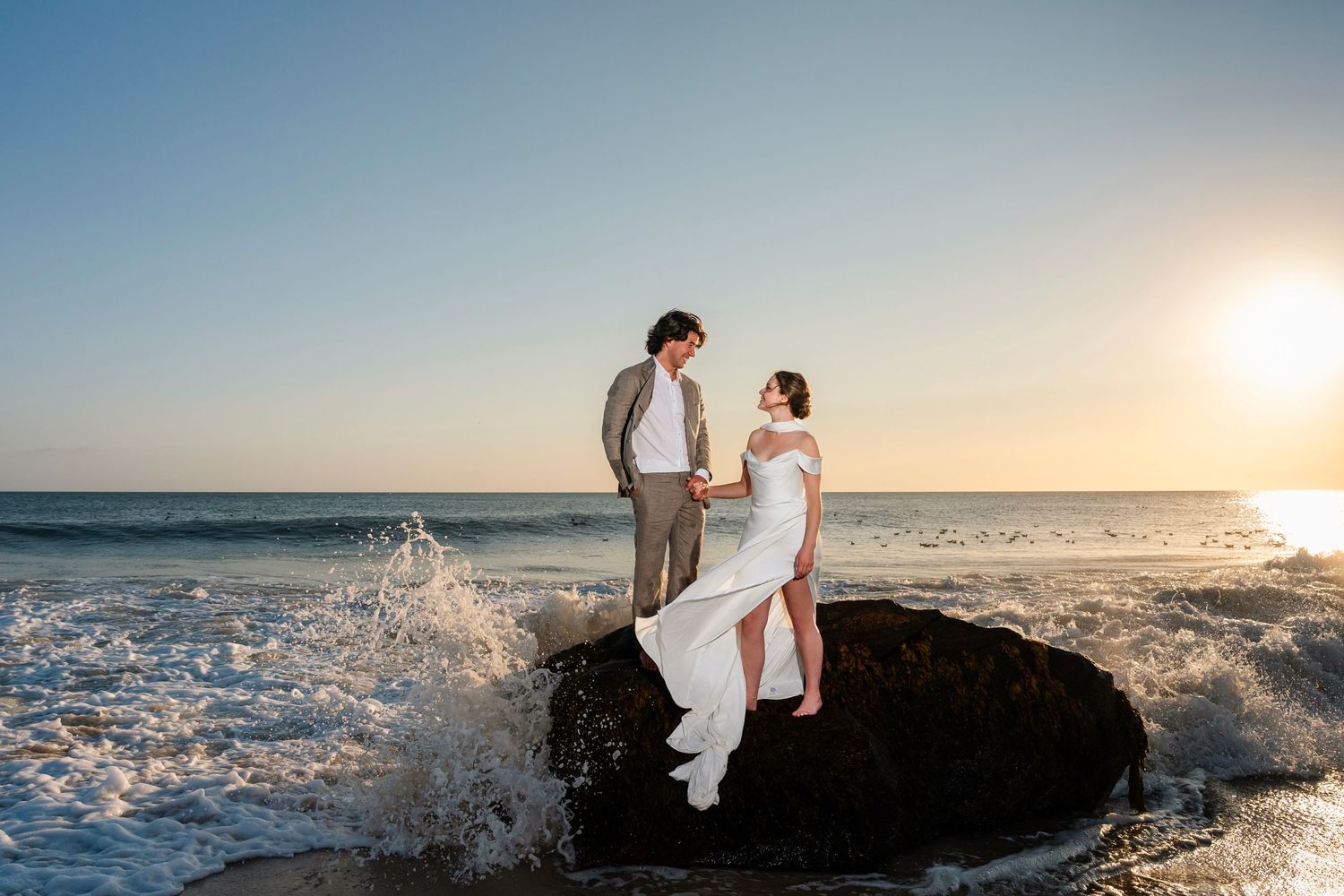 A couple embraces on rocky coastal cliffs as ocean waves crash dramatically at golden hour.