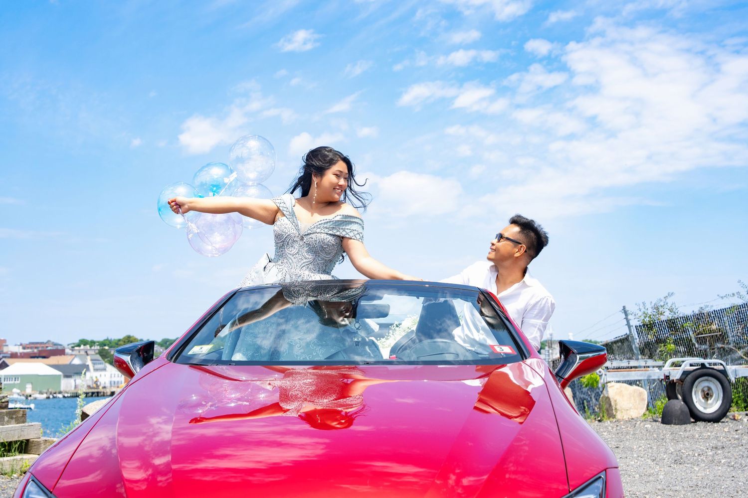 A couple poses playfully with a red convertible car under a bright blue sky.