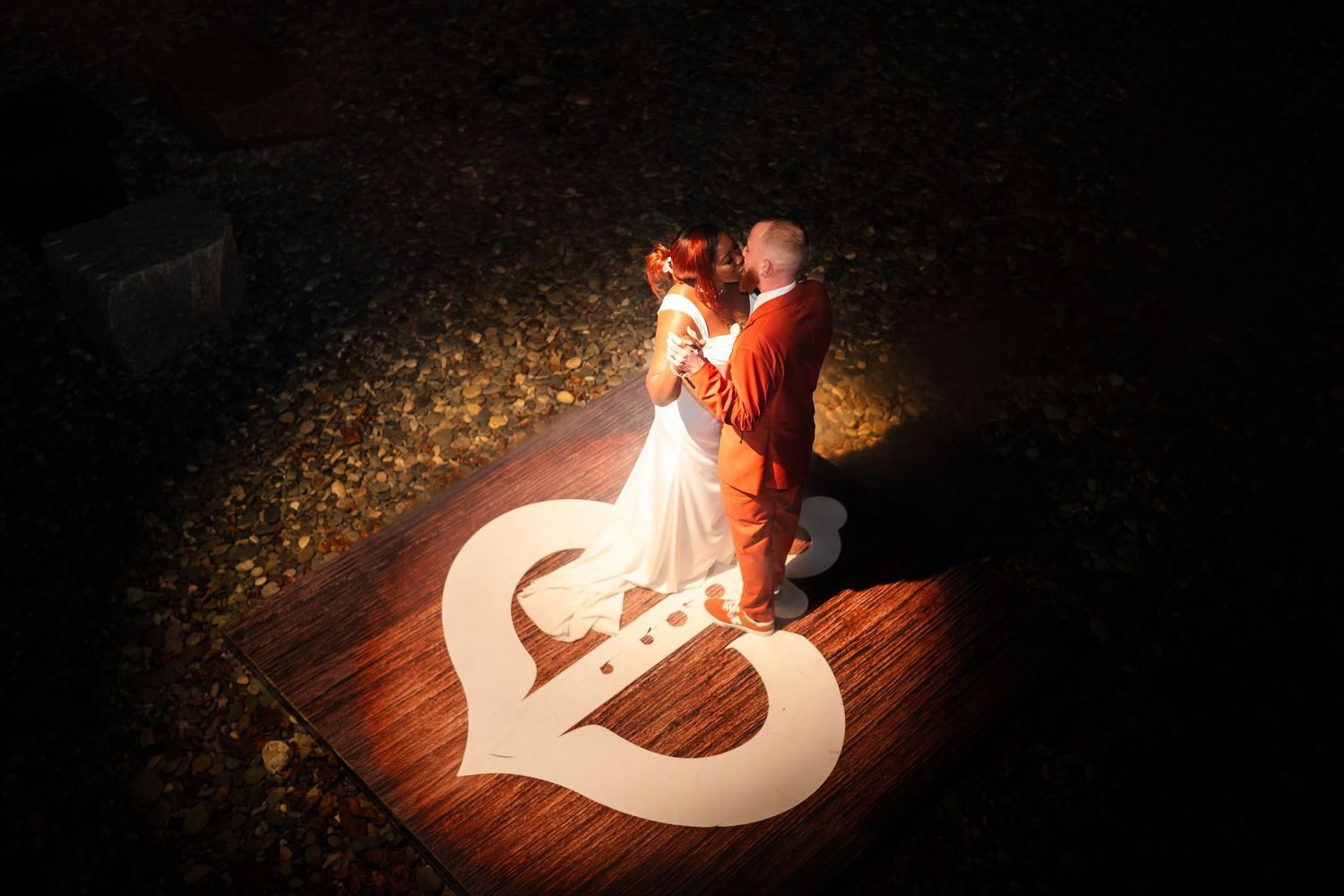 A couple dances on a heart-shaped spotlight on a wooden floor during an evening wedding reception.