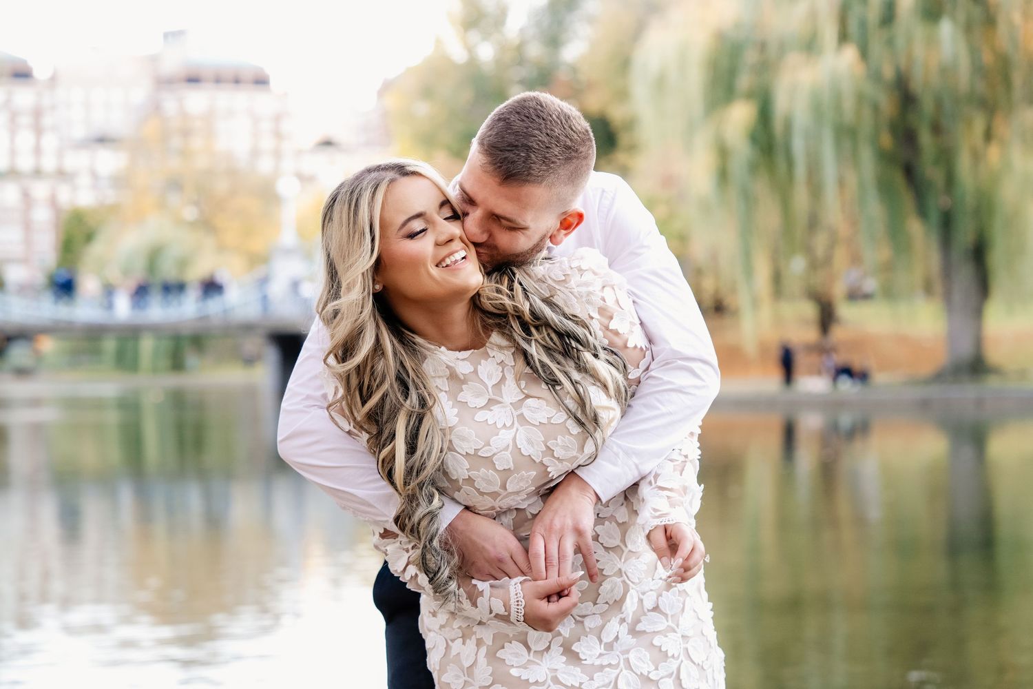 A couple shares an intimate moment near a willow tree beside a tranquil pond in an autumn setting.