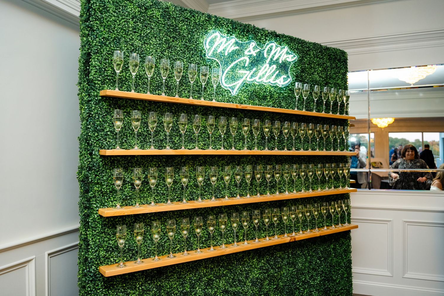 Elegant champagne display wall with green hedge backdrop and wooden shelving at an upscale event venue.
