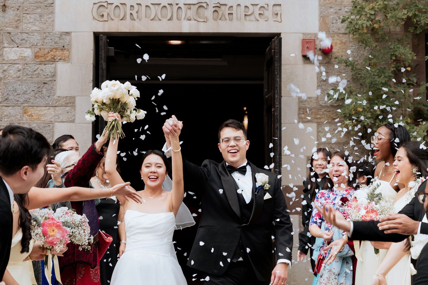 Newlyweds celebrate while guests shower them with white petals outside stone church entrance.