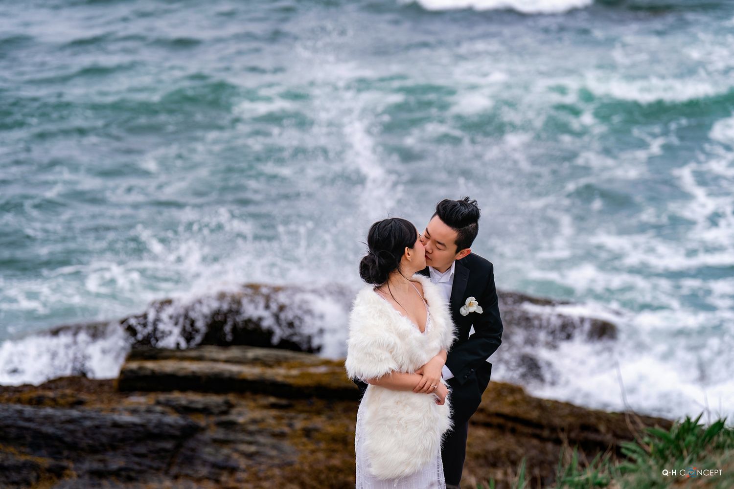 A couple embraces on rocky coastal cliffs with dramatic ocean waves crashing behind them.