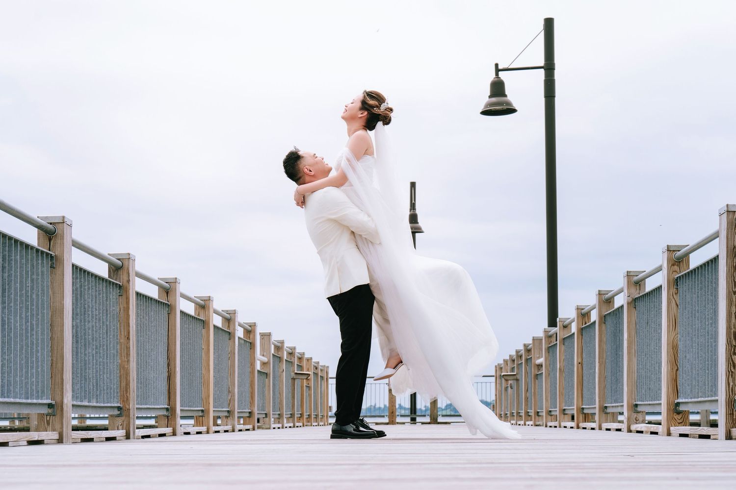 A couple in wedding attire embraces on a wooden pier with a street lamp and cloudy sky in the background.