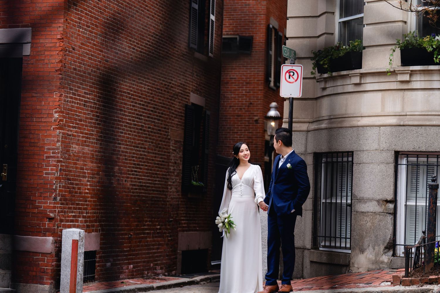 A couple in wedding attire stands together on a brick sidewalk near a historic building in an urban setting.