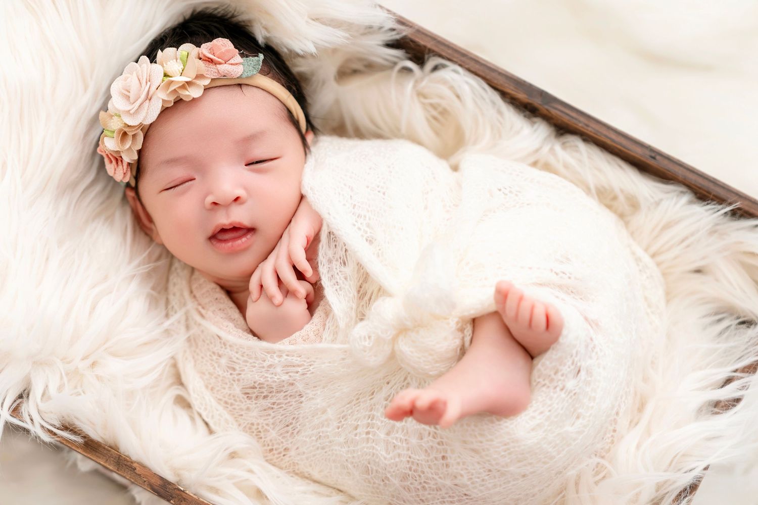 Newborn baby sleeping peacefully in lace wrap with floral headband on fur backdrop.