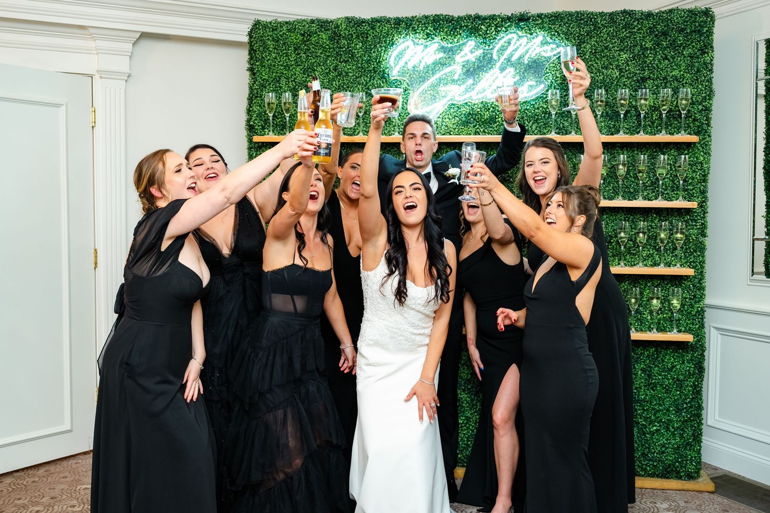 A bridal party in black dresses and one white gown celebrate with champagne bottles against a green wall backdrop.