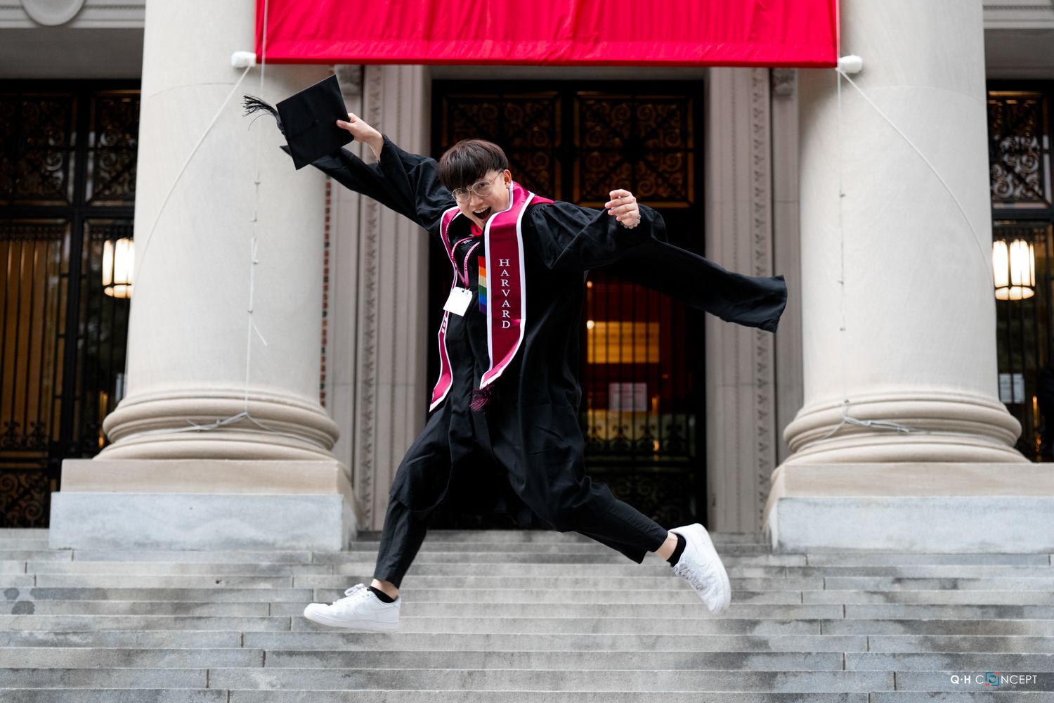 Jubilant graduate in cap and gown leaps on university steps, celebrating commencement with raised fist.
