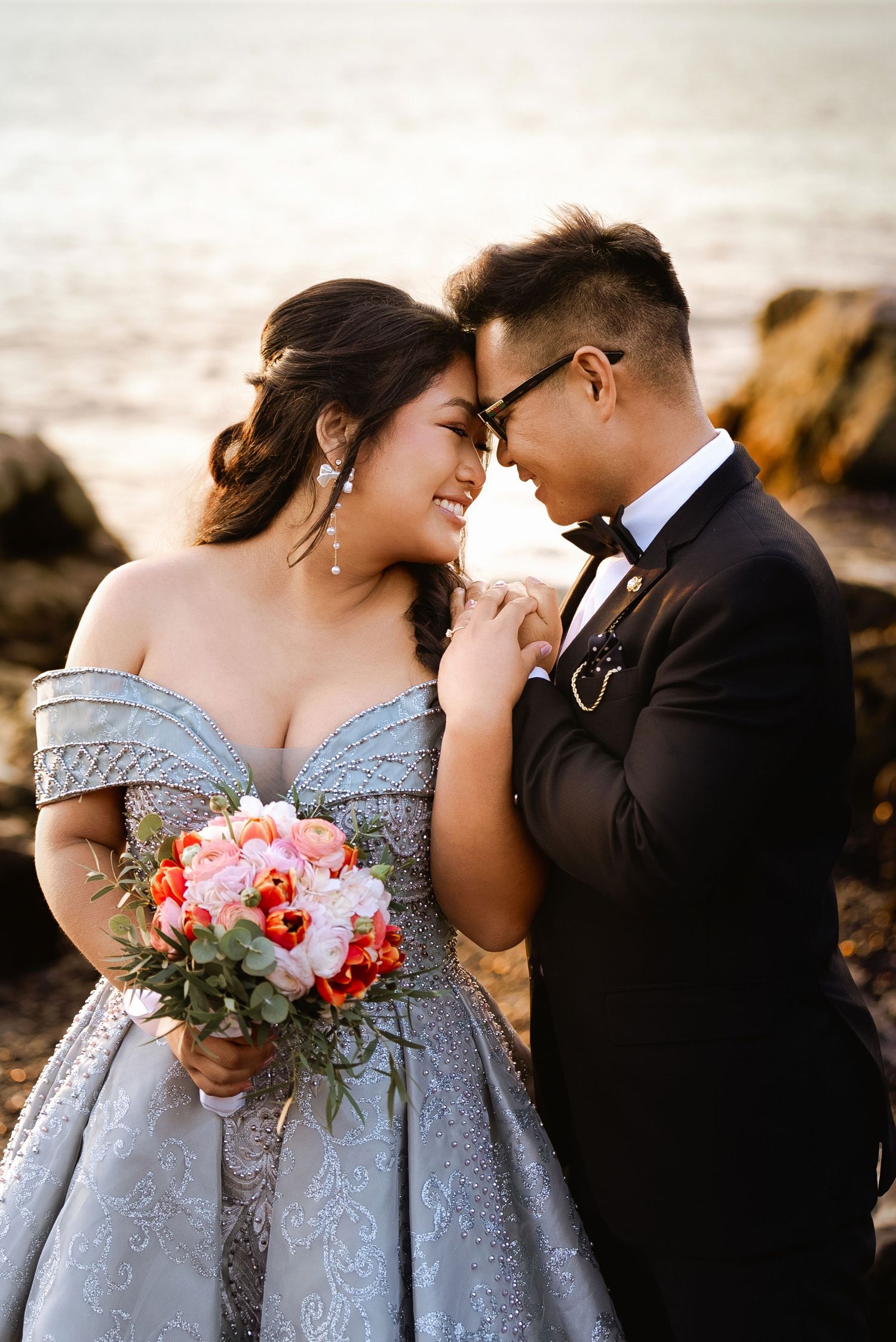 Couple shares tender moments in formal attire by the ocean with coral flowers.