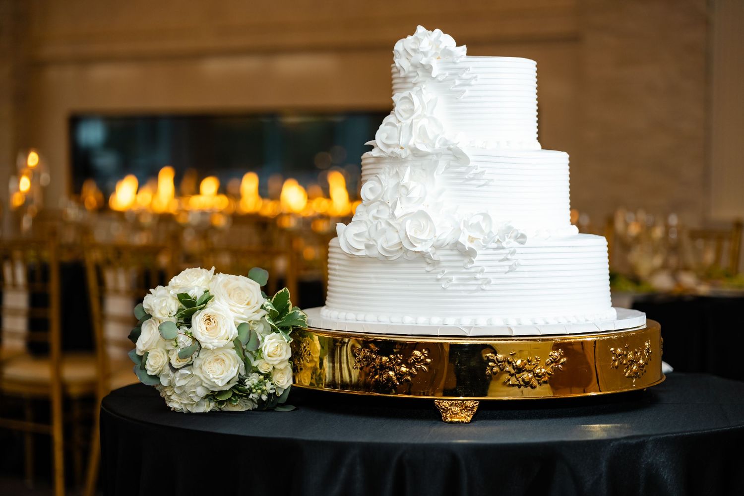 Elegant white tiered wedding cake with floral decorations displayed on a gold stand with a bouquet beside it.
