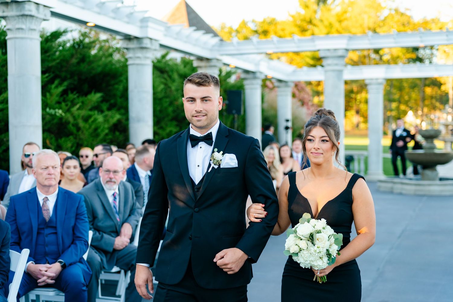 Wedding party members walk down the aisle during an outdoor ceremony under a white pergola with autumn colors in background.