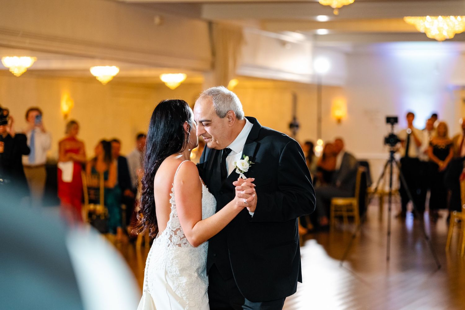 Wedding couple dances together in an elegant ballroom with warm lighting and guests watching.