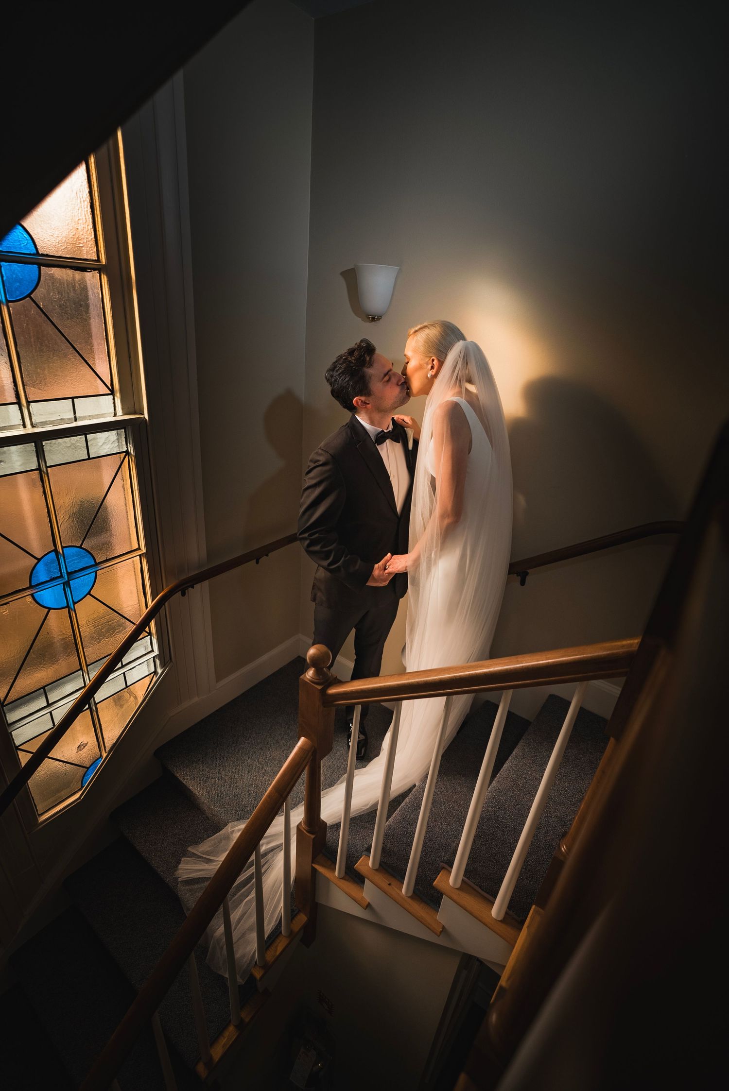 Artistic wedding photo on a staircase with stained glass window and dramatic lighting.