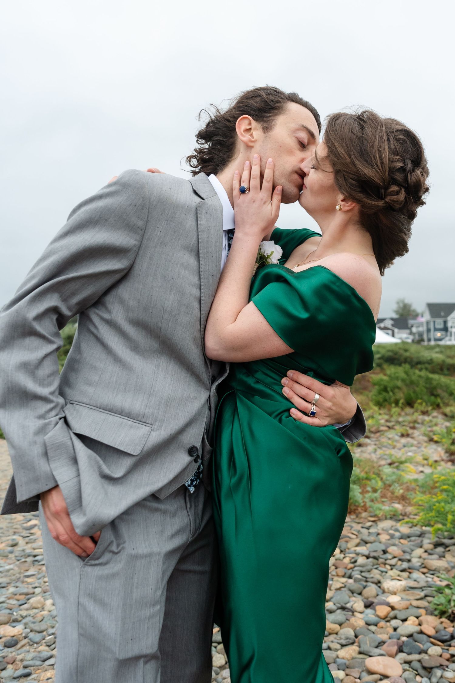 Romantic couple in grey suit and emerald dress share intimate moments on a rocky beach.