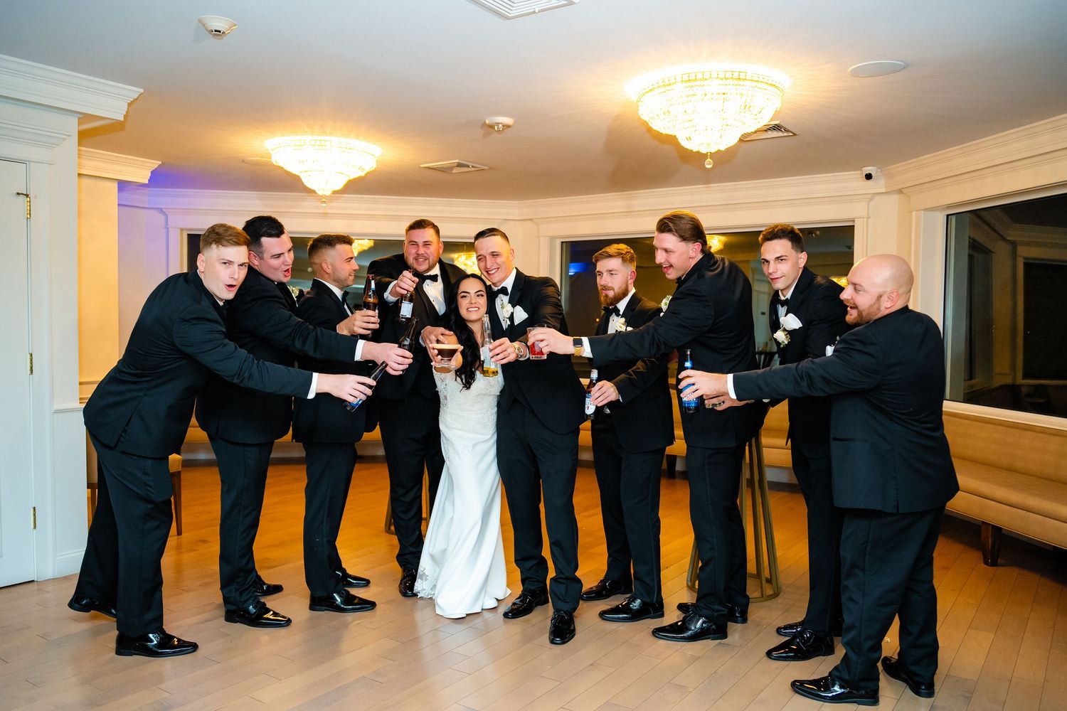 A wedding party in formal attire poses in a line against a light colored wall in a reception venue.