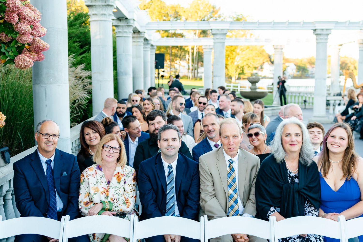 Wedding ceremony guests seated in white chairs on a columned outdoor terrace.