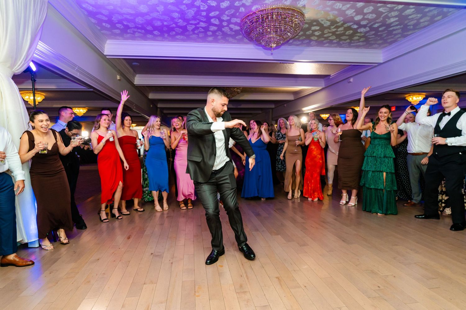 Large group of wedding guests dancing in formation under purple lighting at reception.