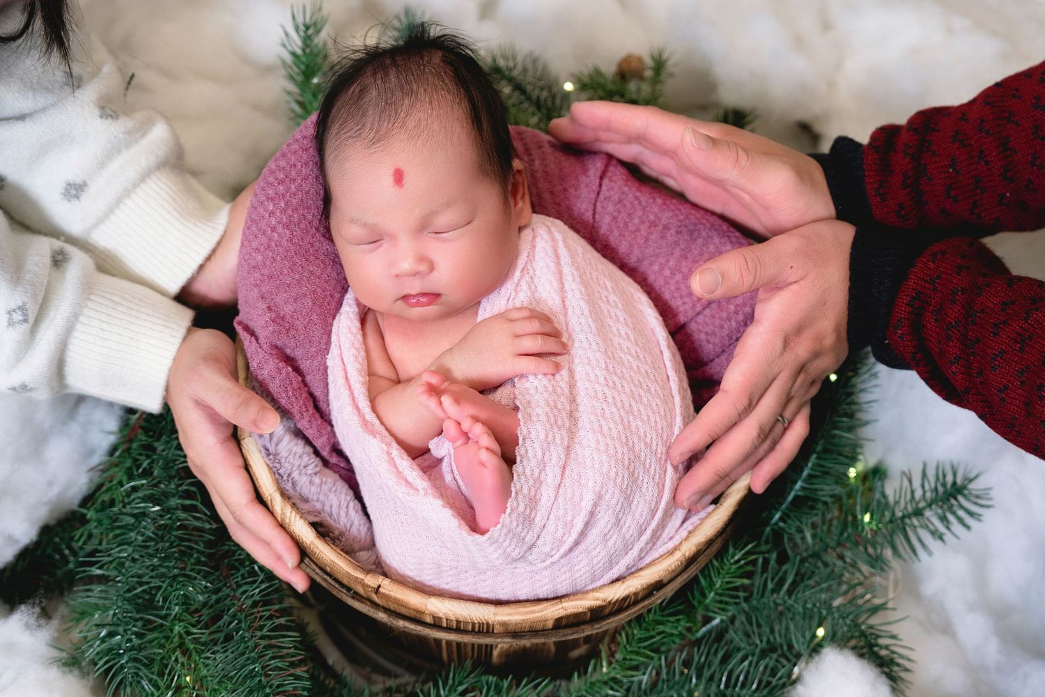 A newborn baby wrapped in pink blanket rests in wooden basket with evergreen branches and gentle hands supporting.