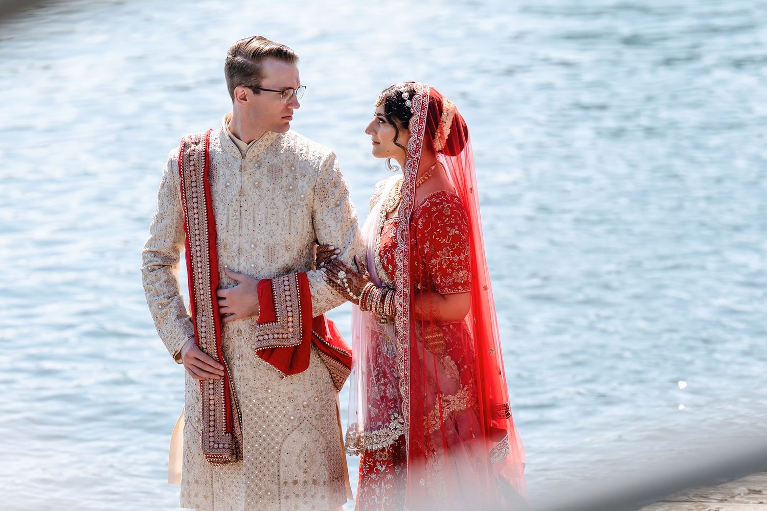 A couple in traditional South Asian wedding attire stands together by a waterfront on their special day.