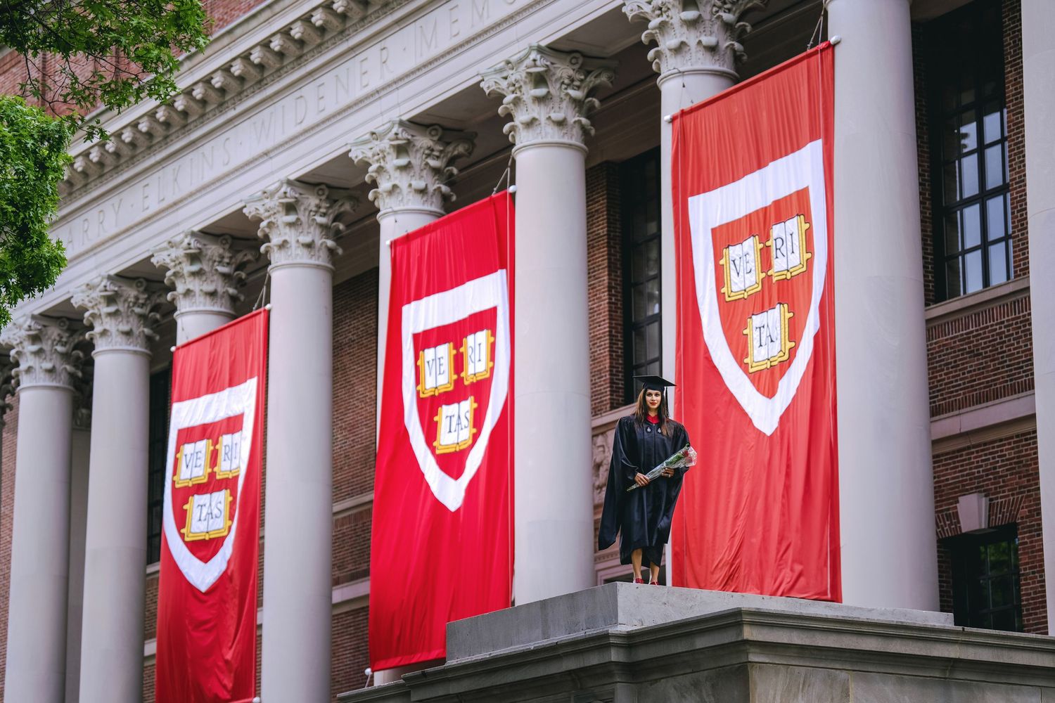 Harvard University building adorned with red banners featuring the school crest, with a graduate in cap and gown standing nearby.