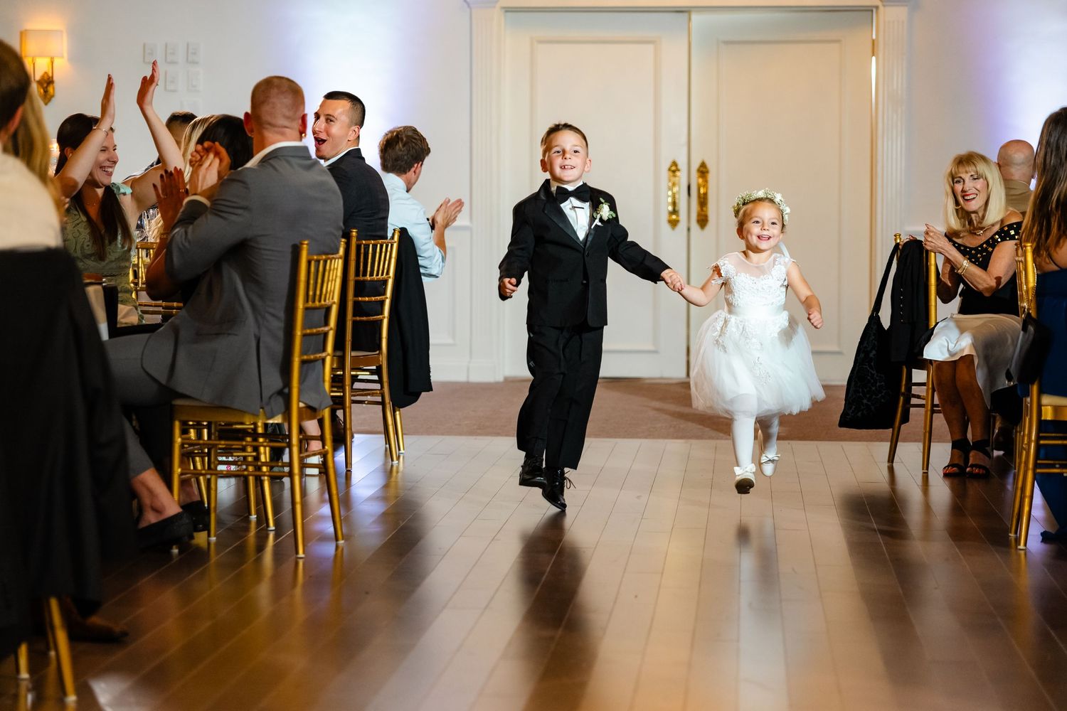 Young ring bearer and flower girl run down aisle at wedding ceremony while guests watch from gold chairs.