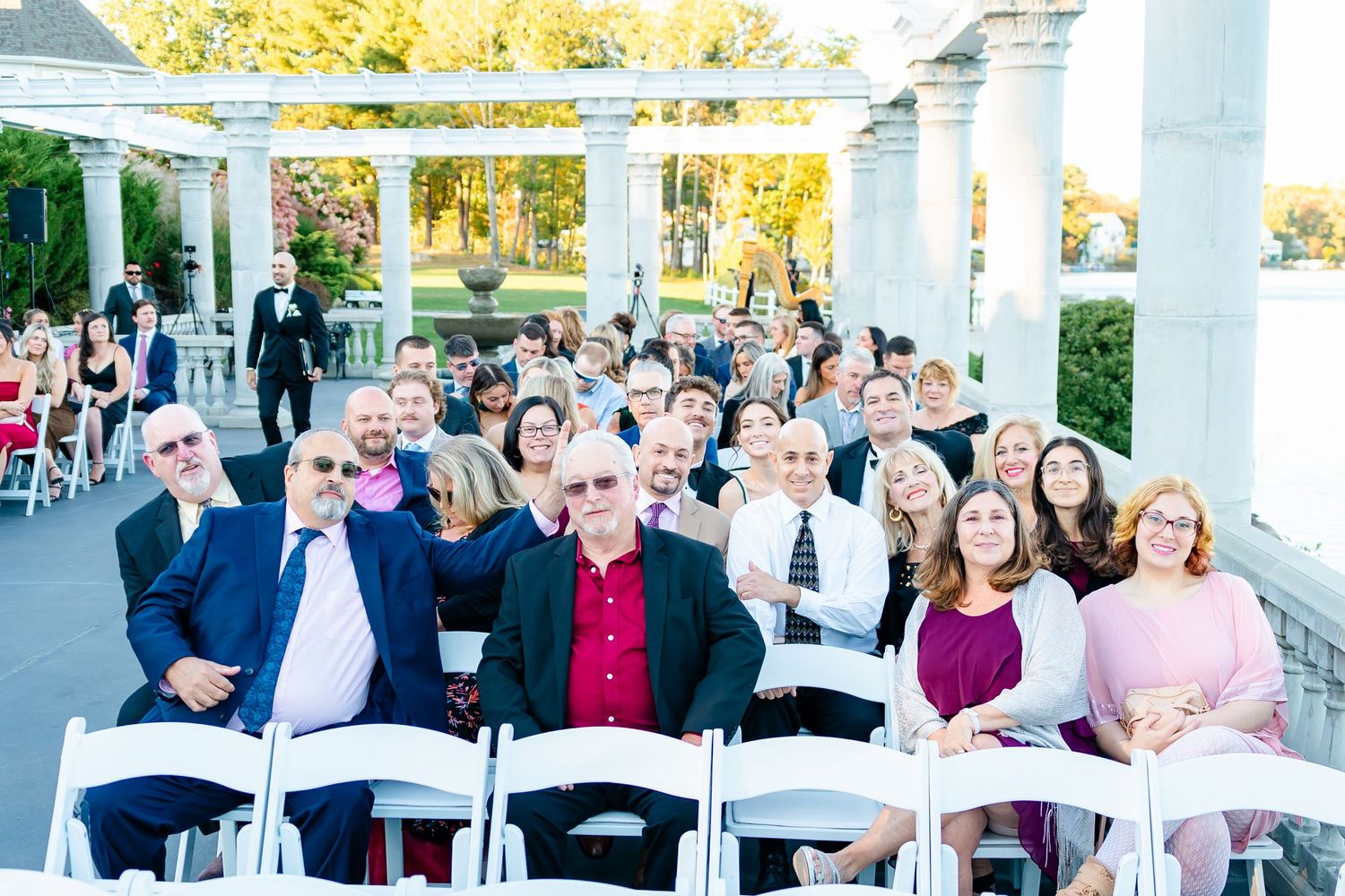 Wedding ceremony guests seated outdoors under white columns with autumn foliage in the background.