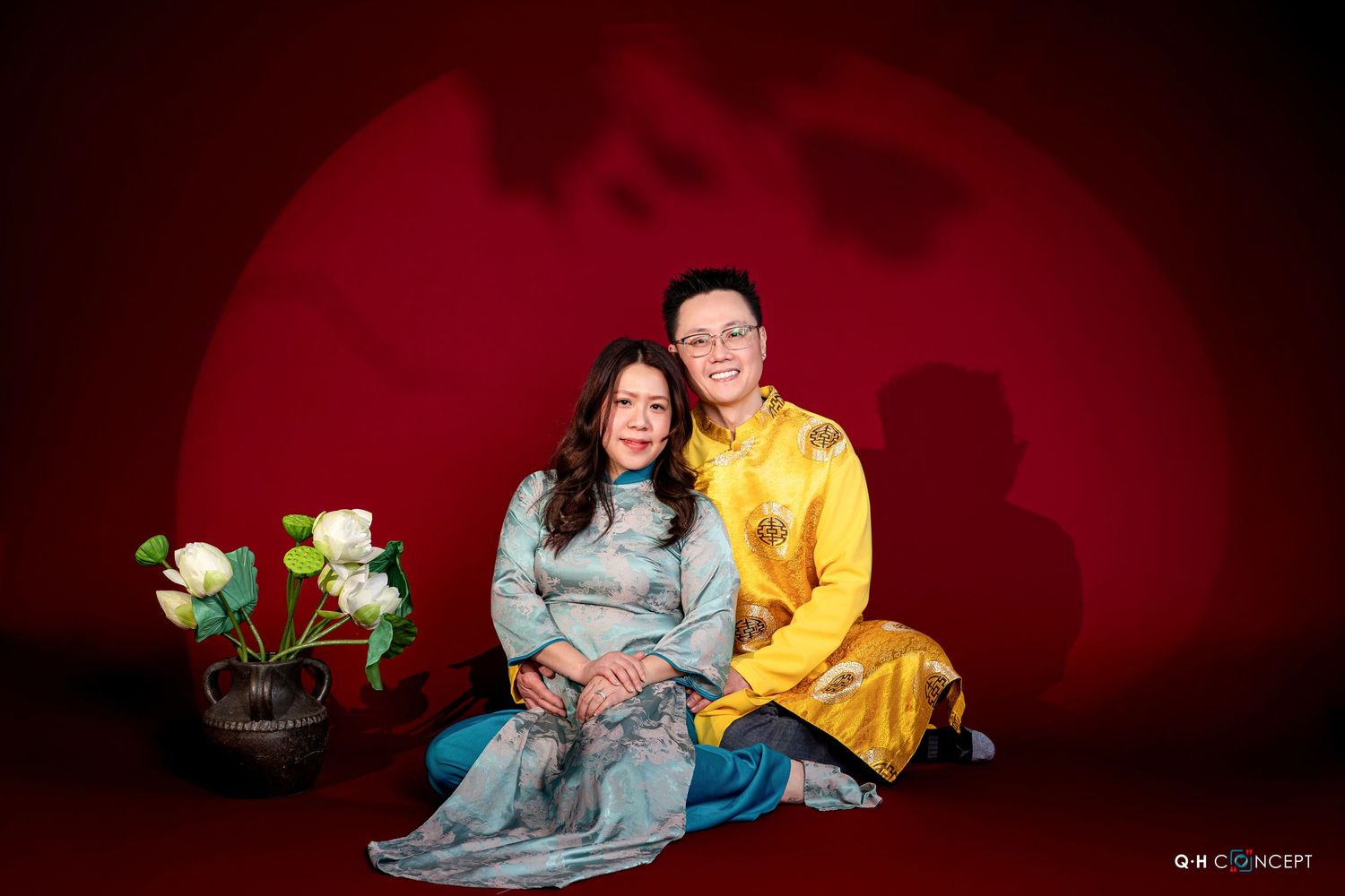 Couple in traditional Asian attire poses lovingly against dark red backdrop in romantic photo session.