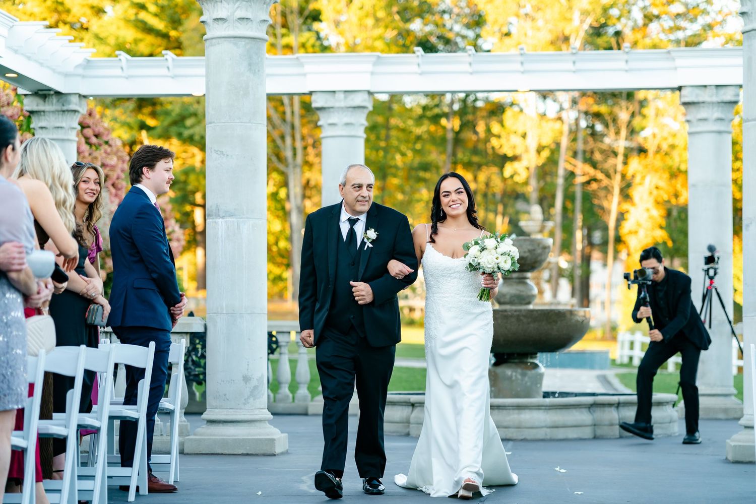 Outdoor wedding ceremony setup with white pillars and chairs arranged in rows beneath golden autumn trees.