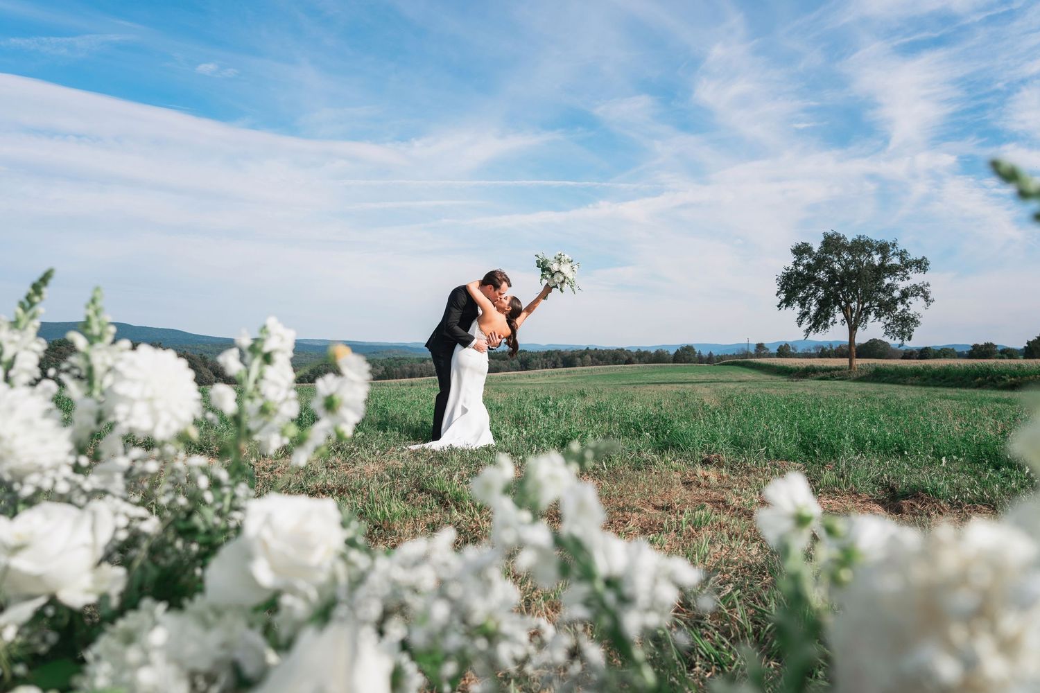 A dreamy outdoor wedding scene with white wildflowers in the foreground and a couple in a green meadow at sunset.